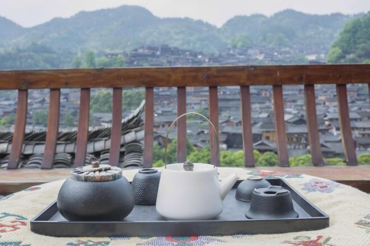 Uma bandeja com uma chaleira branca e utensílios para chá preto sobre uma toalha com padrão tradicional, com vista para uma aldeia com edifícios tradicionais.
