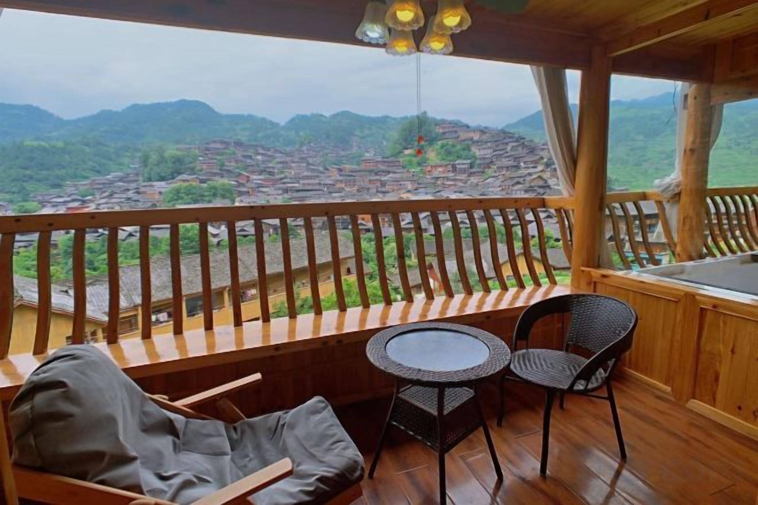 Un balcón de madera con vistas a un pueblo y montañas, una mesa redonda y dos sillas.