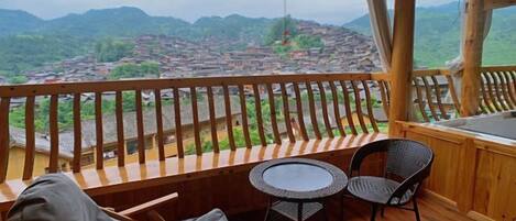 Un balcón de madera con vistas a un pueblo y montañas, una mesa redonda y dos sillas.