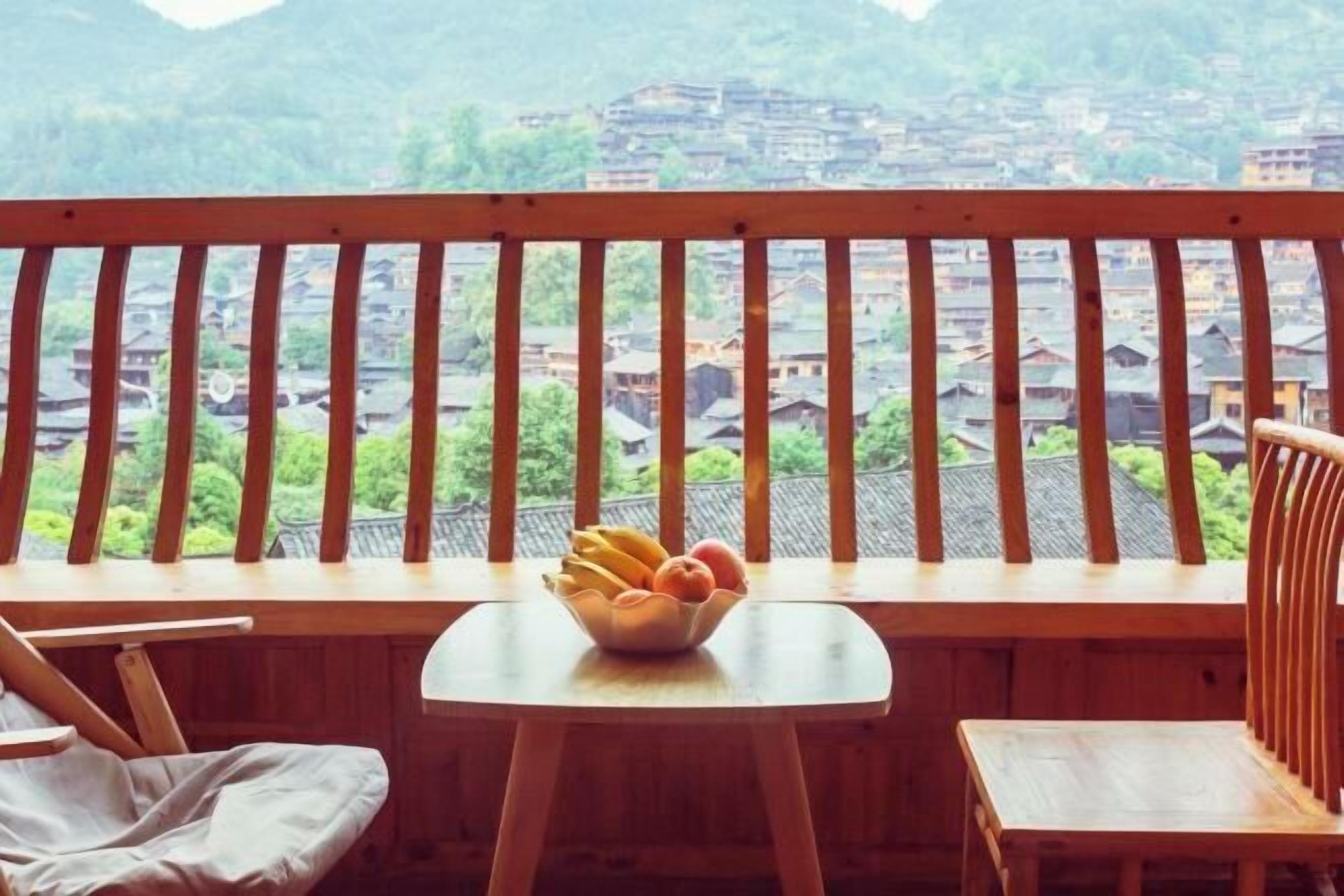 A balcony with a wooden railing, a small table with a bowl of fruit, and a view of traditional buildings in the background.