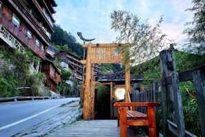 A traditional wooden gate with a sign, a wooden bench, and a road leading to a building with a sloped roof.
