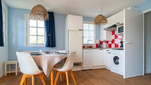 A compact kitchen with a washing machine, a dining area with a table and chairs, and a window with curtains. - Côté Ouest - Appt avec piscine partagée (Benodet)