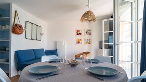 A dining area with a round table set for four, a blue sofa, and a basket hanging on the wall. - Côté Ouest - Appt avec piscine partagée (Benodet)