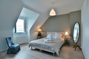 Un dormitorio con techo inclinado, una cama con ropa de cama blanca, dos lámparas de noche, un espejo grande y una silla.