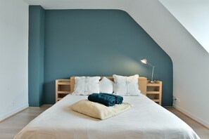 Un dormitorio con techo inclinado, una cama con ropa de cama blanca, una toalla azul y una lámpara de noche.