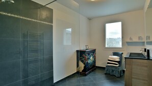 A modern bathroom with a glass shower enclosure, a small seating area with a towel stack, and a window providing natural light. - Côté Plage - Maison  pour 12 vue mer (Saint-Cast-le-Guildo)