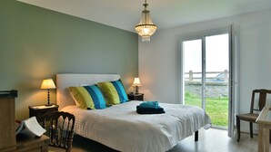 A bedroom with a large bed, two bedside lamps, a wooden chair, a desk with a chair, and a view of the outdoors through a glass door. - Côté Plage - Maison  pour 12 vue mer (Saint-Cast-le-Guildo)