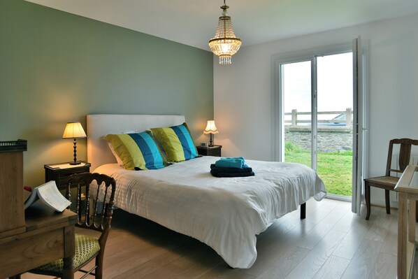 A bedroom with a large bed, two bedside lamps, a wooden chair, a desk with a chair, and a view of the outdoors through a glass door.