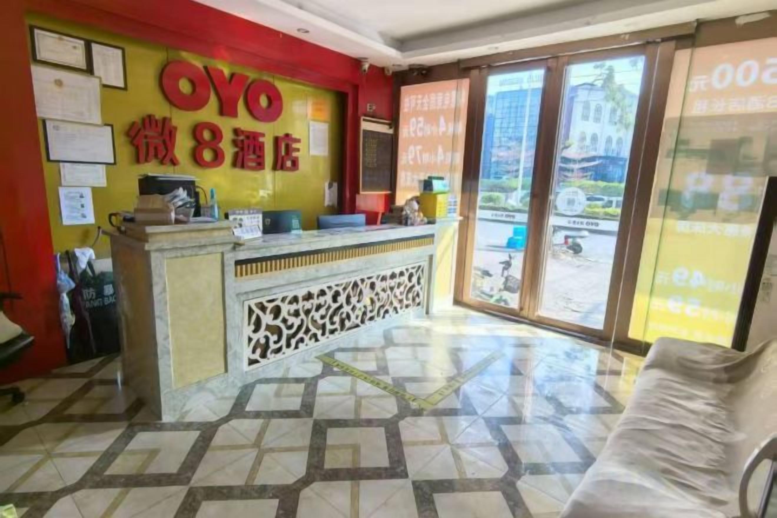 A hotel reception area with a reception desk, a chair, and a large window offering a view of the street.