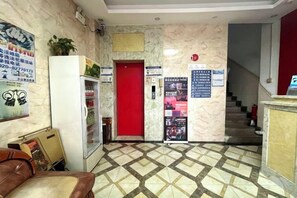 Un vestíbulo de hotel con una recepción, una puerta roja, una escalera y un refrigerador.