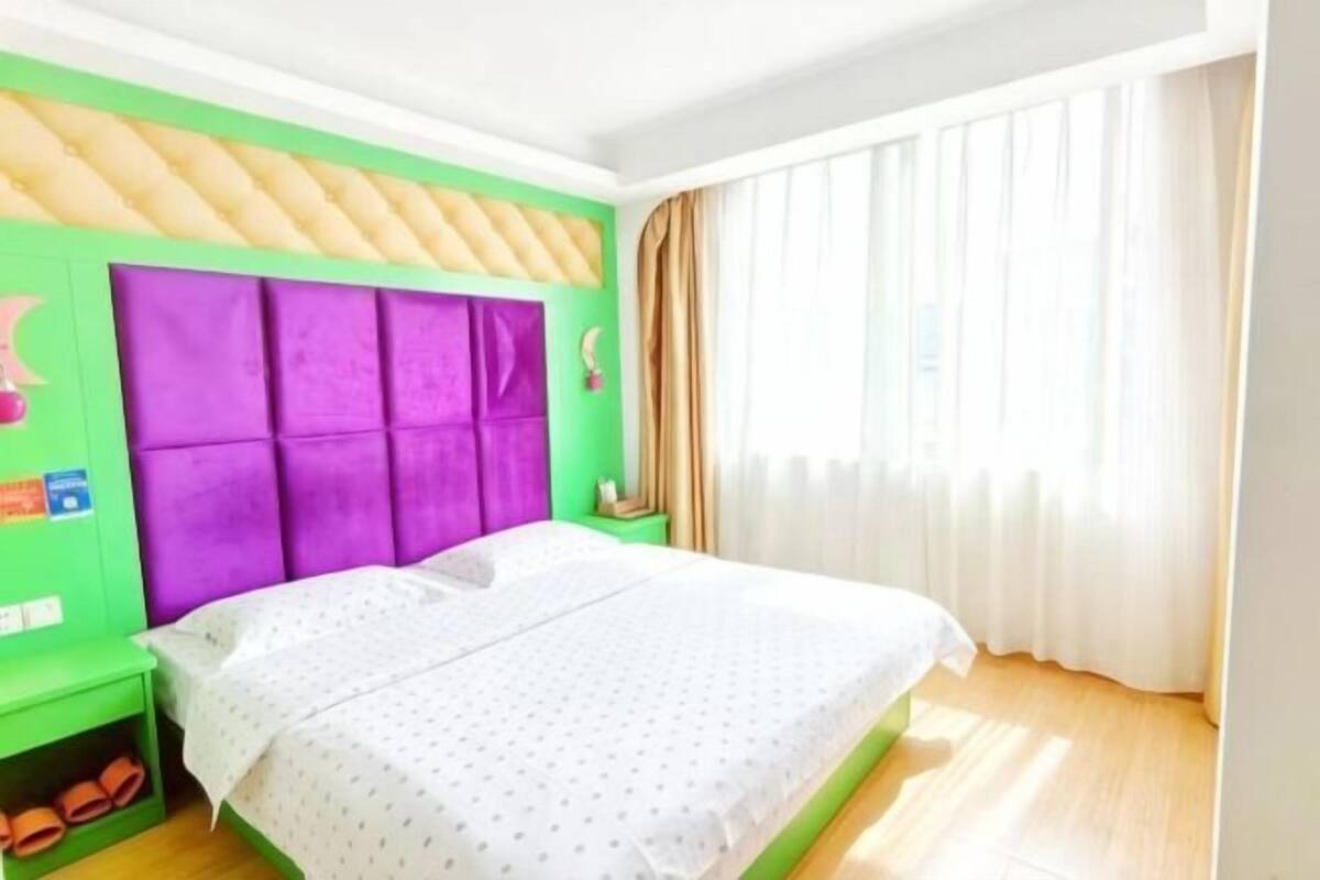 A bedroom with a green wall, a purple headboard, a white bedspread, and a window with curtains.
