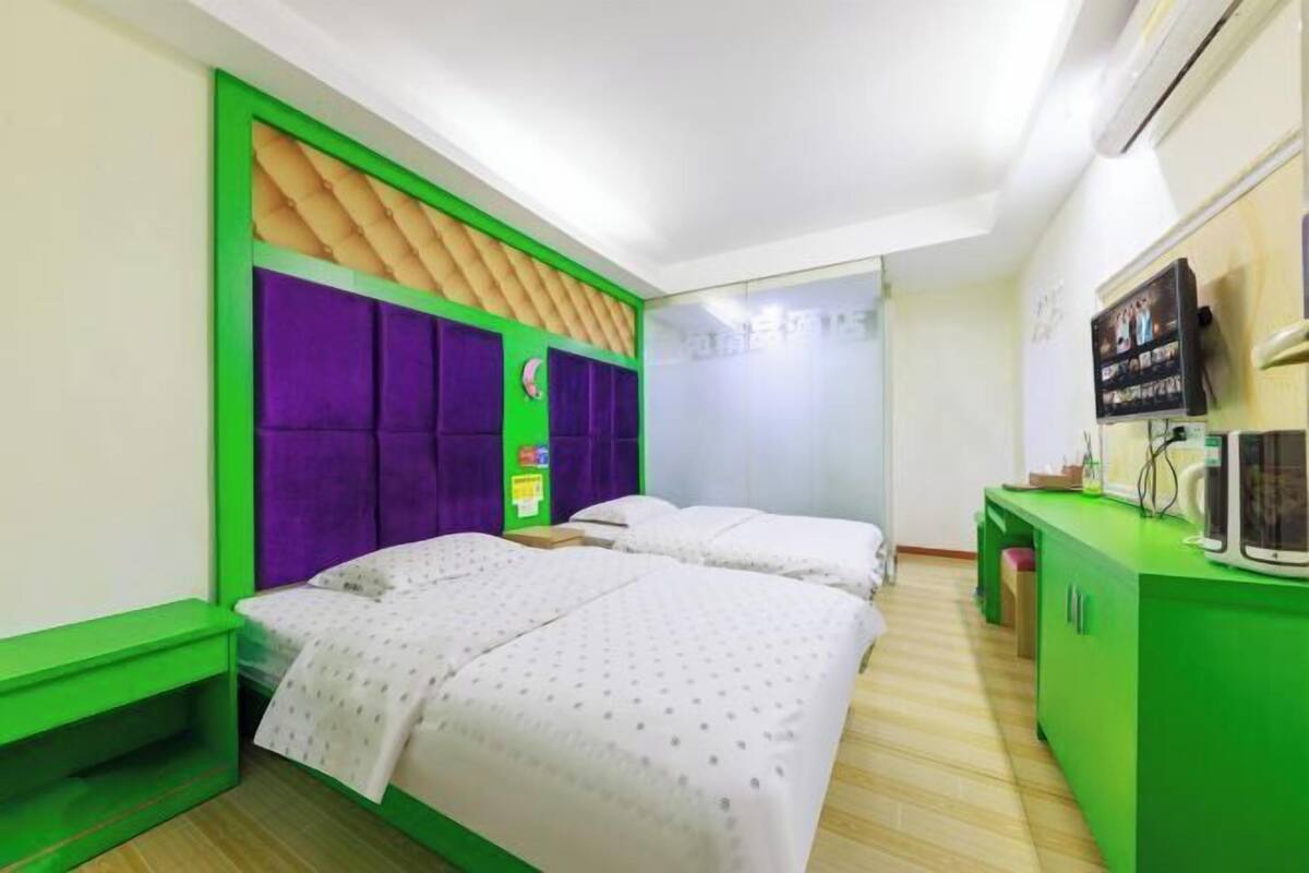 A room with two beds, a green desk, and a television on the wall.