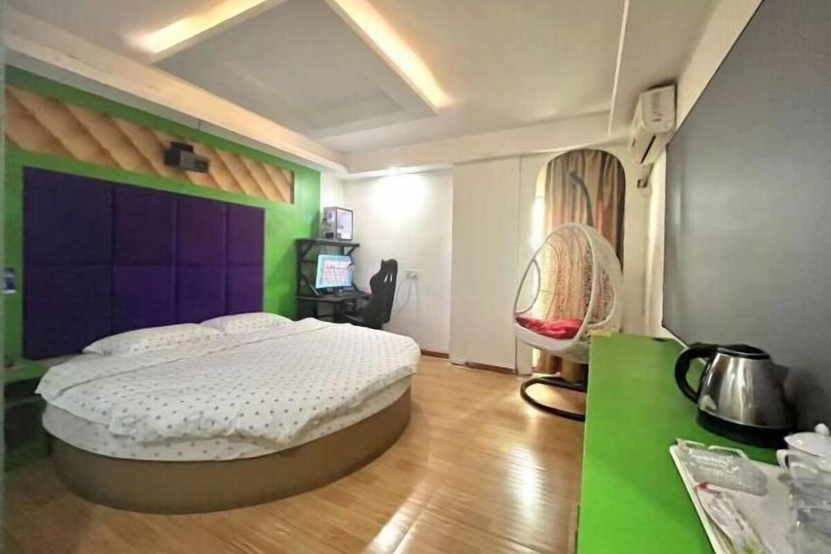 A modern room with a curved bed, a green accent wall, a purple headboard, a small desk with a computer, and a hanging chair.
