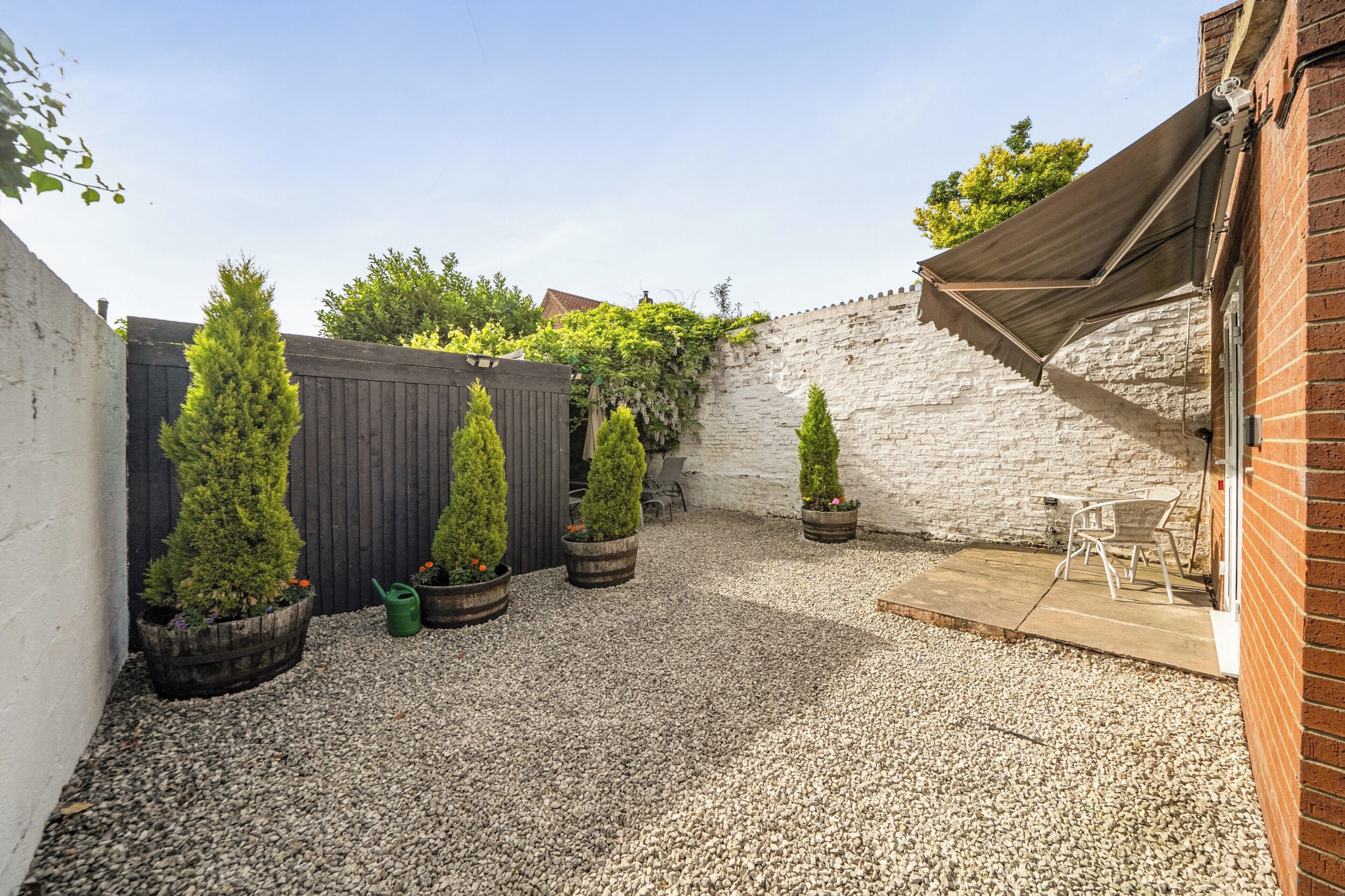 A courtyard with a gravel ground, a wooden fence, potted coniferous trees, a brick wall, and a patio area with outdoor furniture.