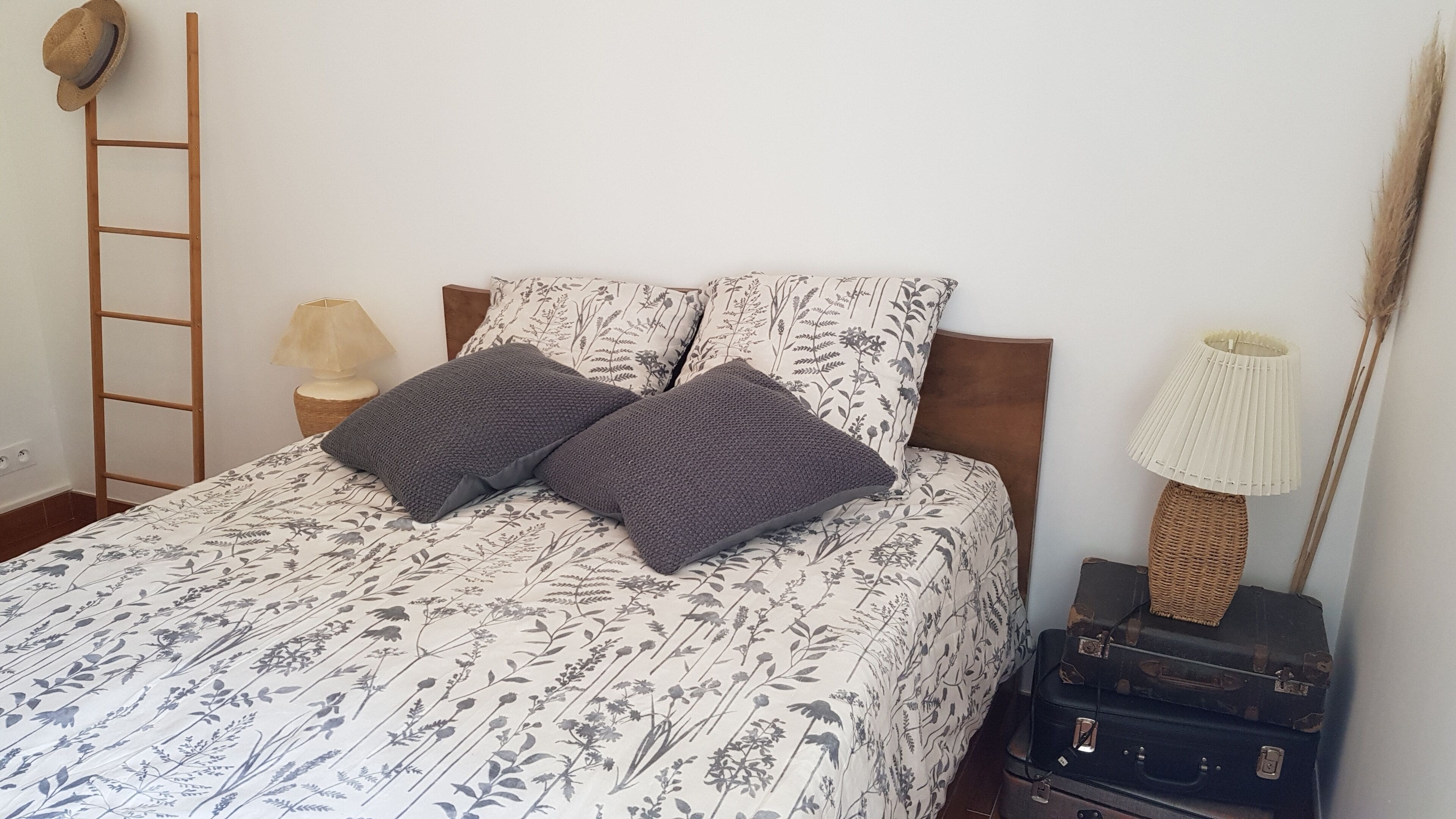 Un lit avec une literie à motifs floraux, deux oreillers gris, une échelle en bois, une lampe et une pile de bagages.