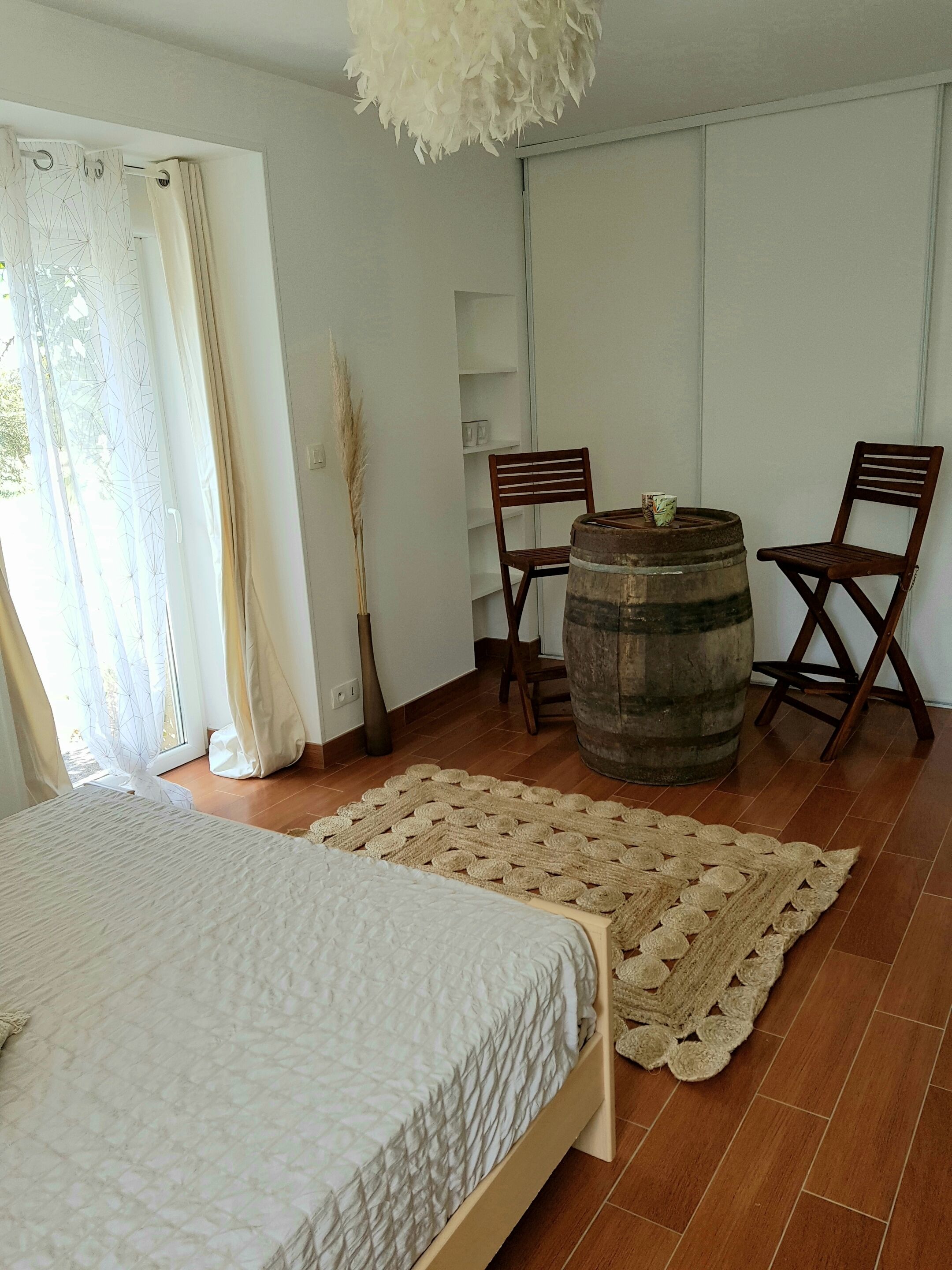 Un dormitorio con una cama, un barril de madera, dos sillas, un tapete tejido y una planta alta.