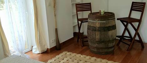 Un dormitorio con una cama, un barril de madera, dos sillas, un tapete tejido y una planta alta.