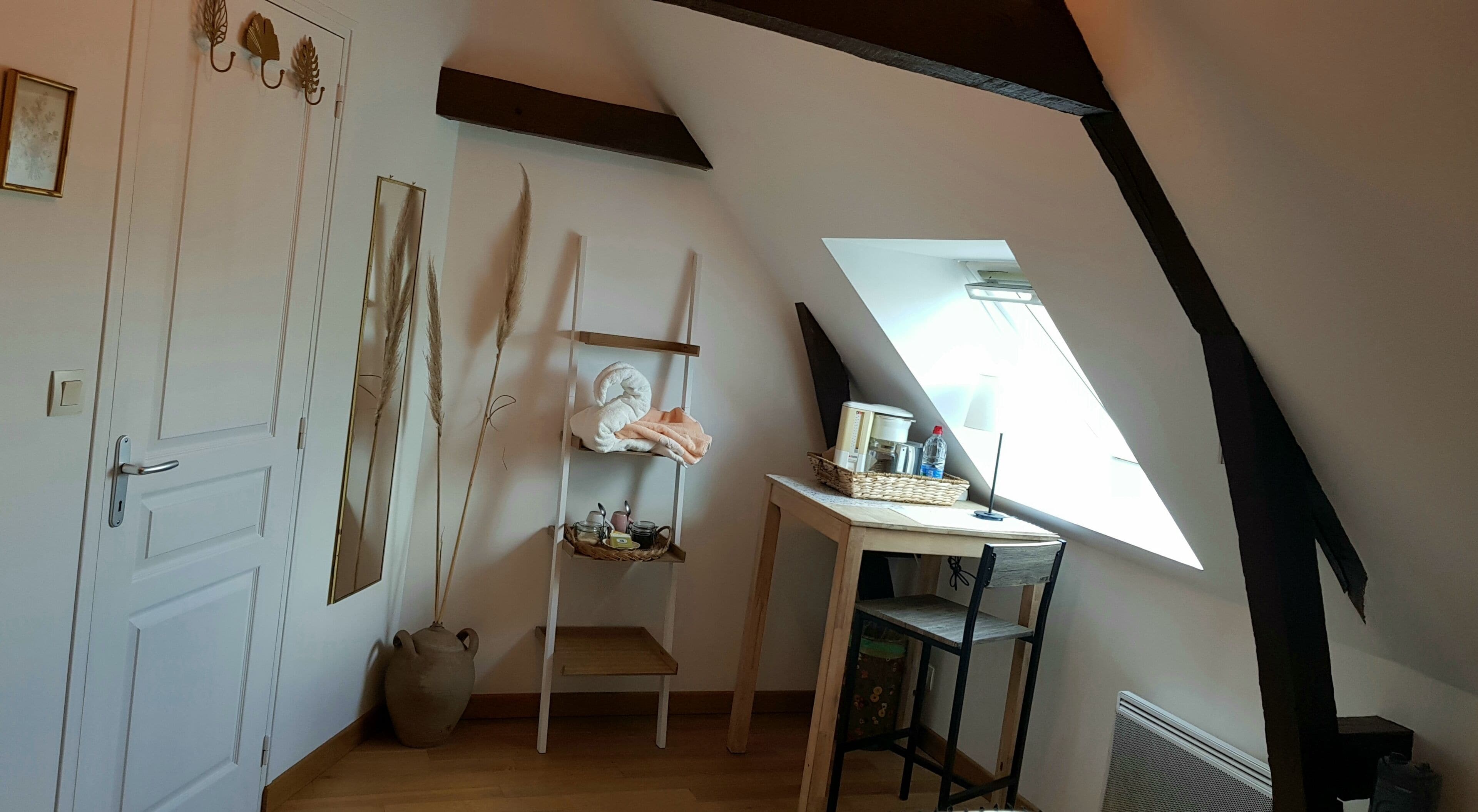 A loft bedroom with a sloped ceiling, a small table with a basket, a shelf with towels, and a door with a mirror.