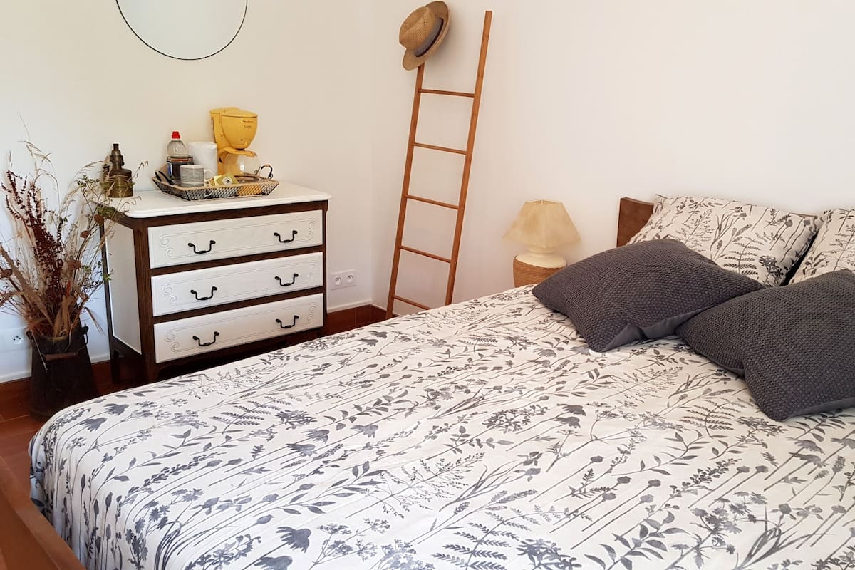 A bedroom with a floral-patterned bedspread, a bedside table with a lamp, a dresser with drawers, and a ladder against the wall.