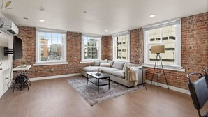 Amplia sala de estar con pared de ladrillo, ventanas grandes, un sofá, una mesa de centro y una lámpara de pie.