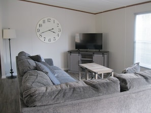 A living room with a grey sofa, a TV on a wooden stand, and a clock on the wall.
