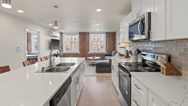 A modern kitchen with white cabinetry, a large island with a sink, and a built-in oven. There is a brick wall and large windows providing natural light. - King Bed! J & K on Main Luxury Apartment 302 (Spokane)