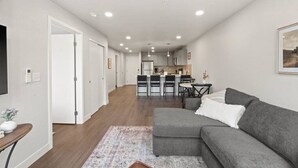 Un apartamento moderno con un sofá seccional gris, una pequeña mesa de comedor y una cocina al fondo.