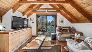 A cozy attic bedroom with a wooden ceiling, a large window, a television, a sofa, and a small table. - Aarlaur 2 - Charming 2BR Rustic Home l Bath l Fire Pit l WiFi Air-Con (Jindabyne)