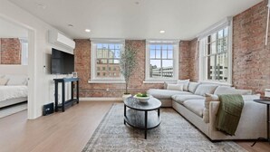 A modern living room with a brick wall, a large window, a sofa, a TV, and a coffee table. - Spacious Apartment with King Beds!  J&K on Main (Spokane)