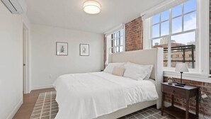 A bedroom with a large bed, a nightstand, a lamp, and a window with a view of buildings. - Spacious Apartment with King Beds!  J&K on Main (Spokane)