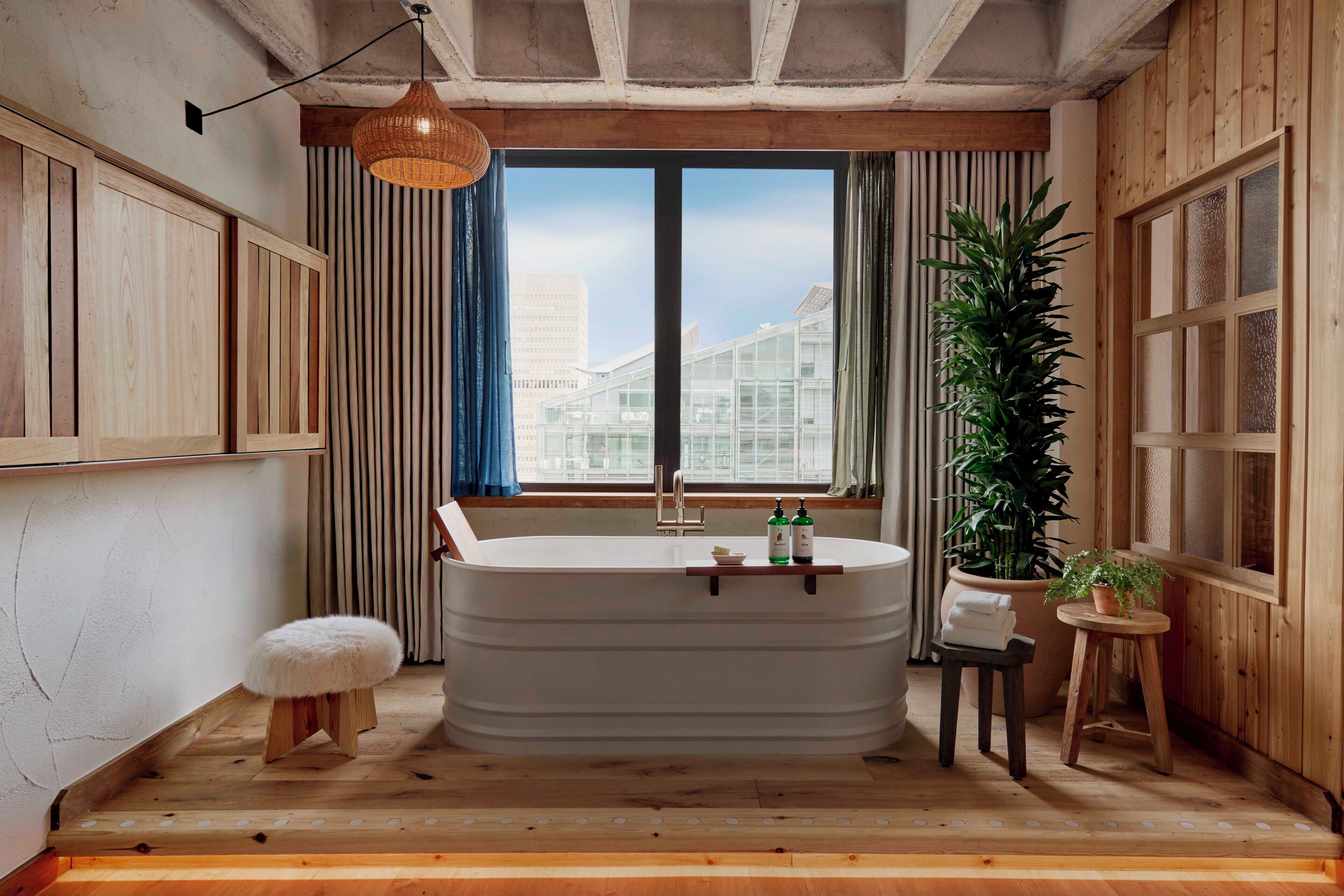 A modern bathroom with a freestanding bathtub, wooden paneling, a large window with a view of buildings, and a hanging light fixture.