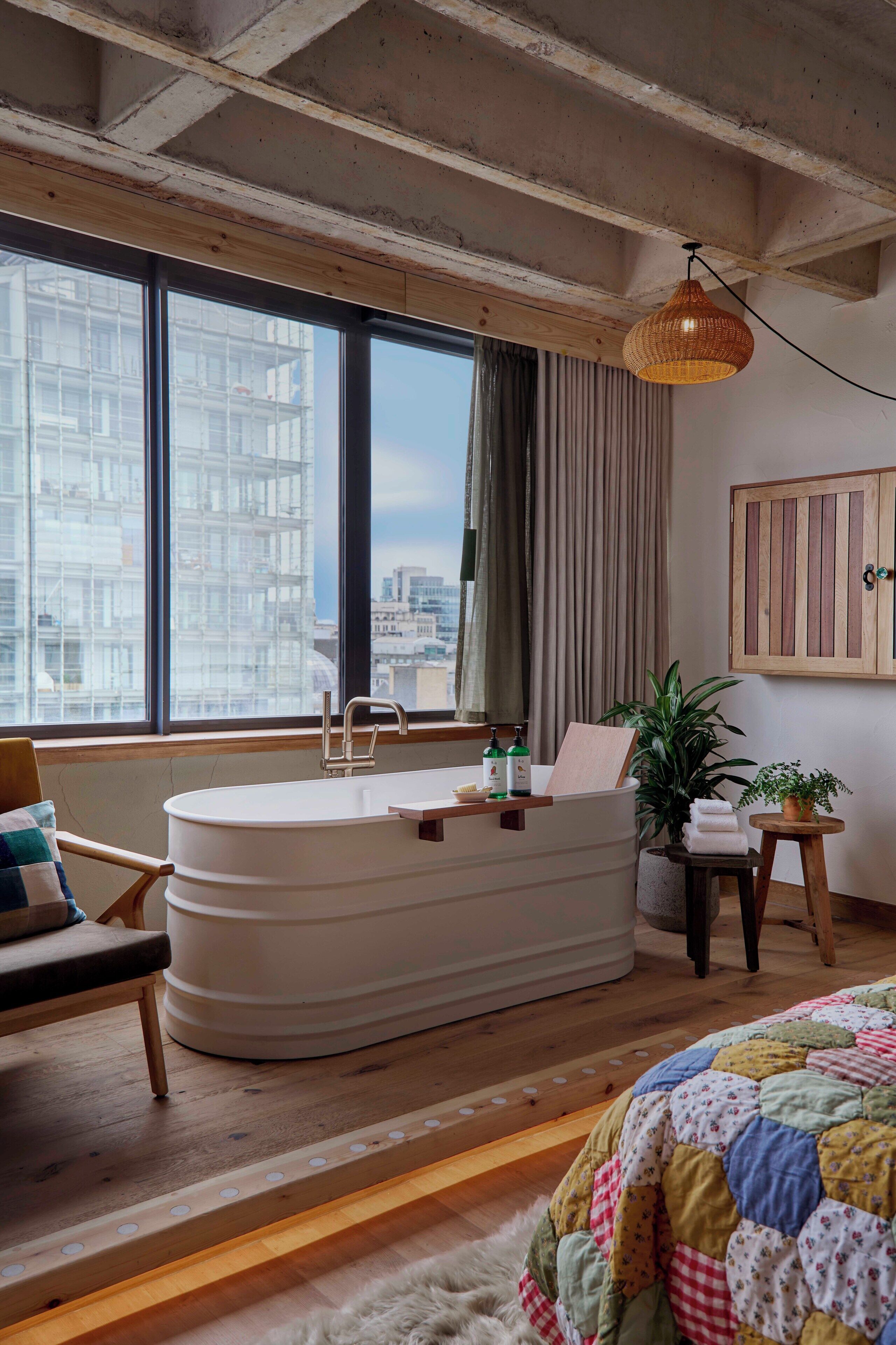 A modern bathroom with a freestanding bathtub, a window with a view of buildings, a wooden chair, a small table with toiletries, and a hanging light fixture.