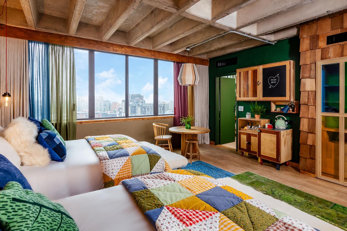 A modern room with a large window, a bed with a colorful quilt, a small table with chairs, and a wooden cabinet with a chalkboard.