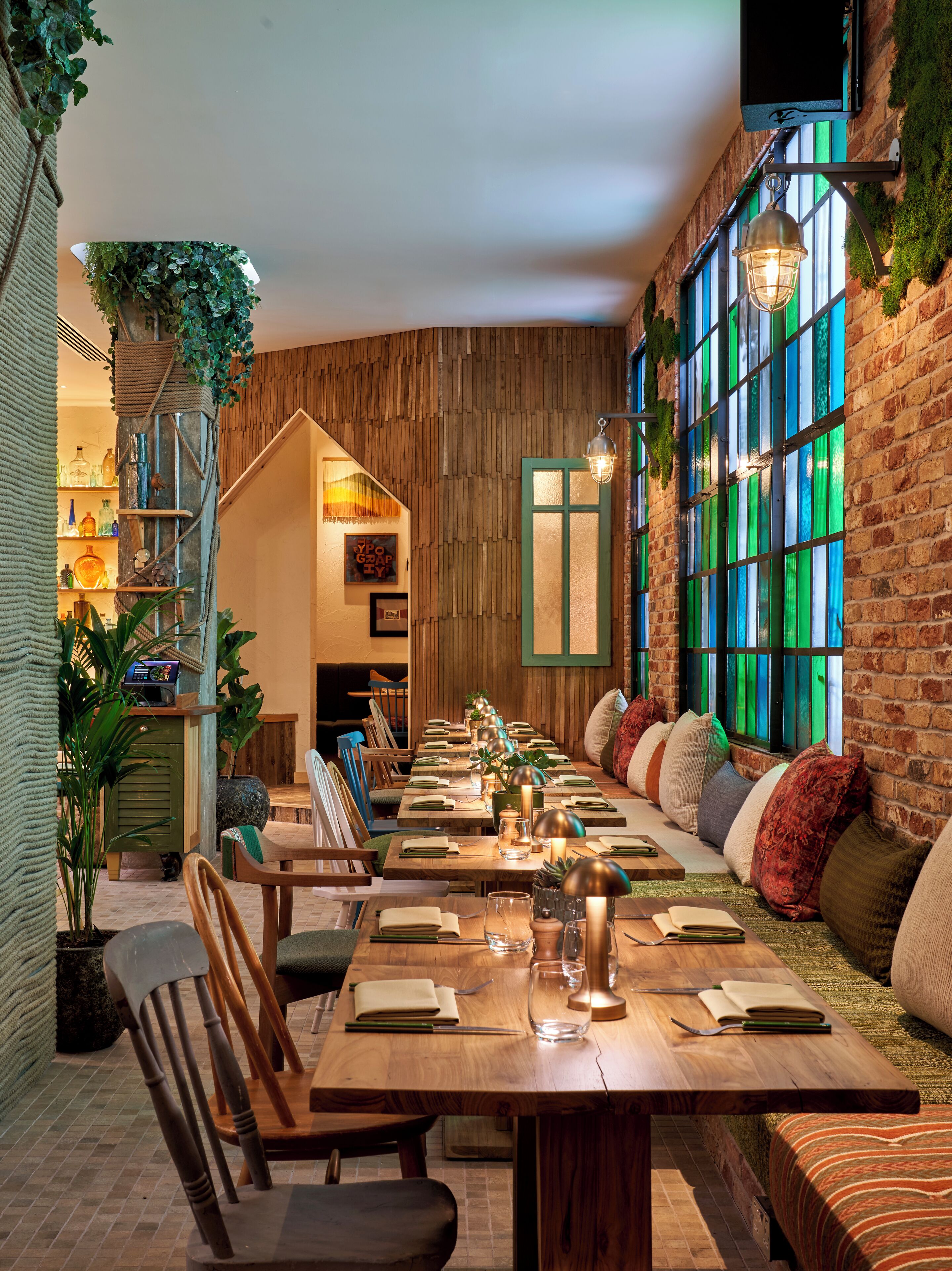 A cozy restaurant with a long wooden table set for dining, surrounded by comfortable seating and a brick wall with green-tinted windows.