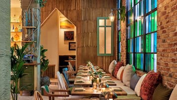 A cozy restaurant with a long wooden table set for dining, surrounded by comfortable seating and a brick wall with green-tinted windows.