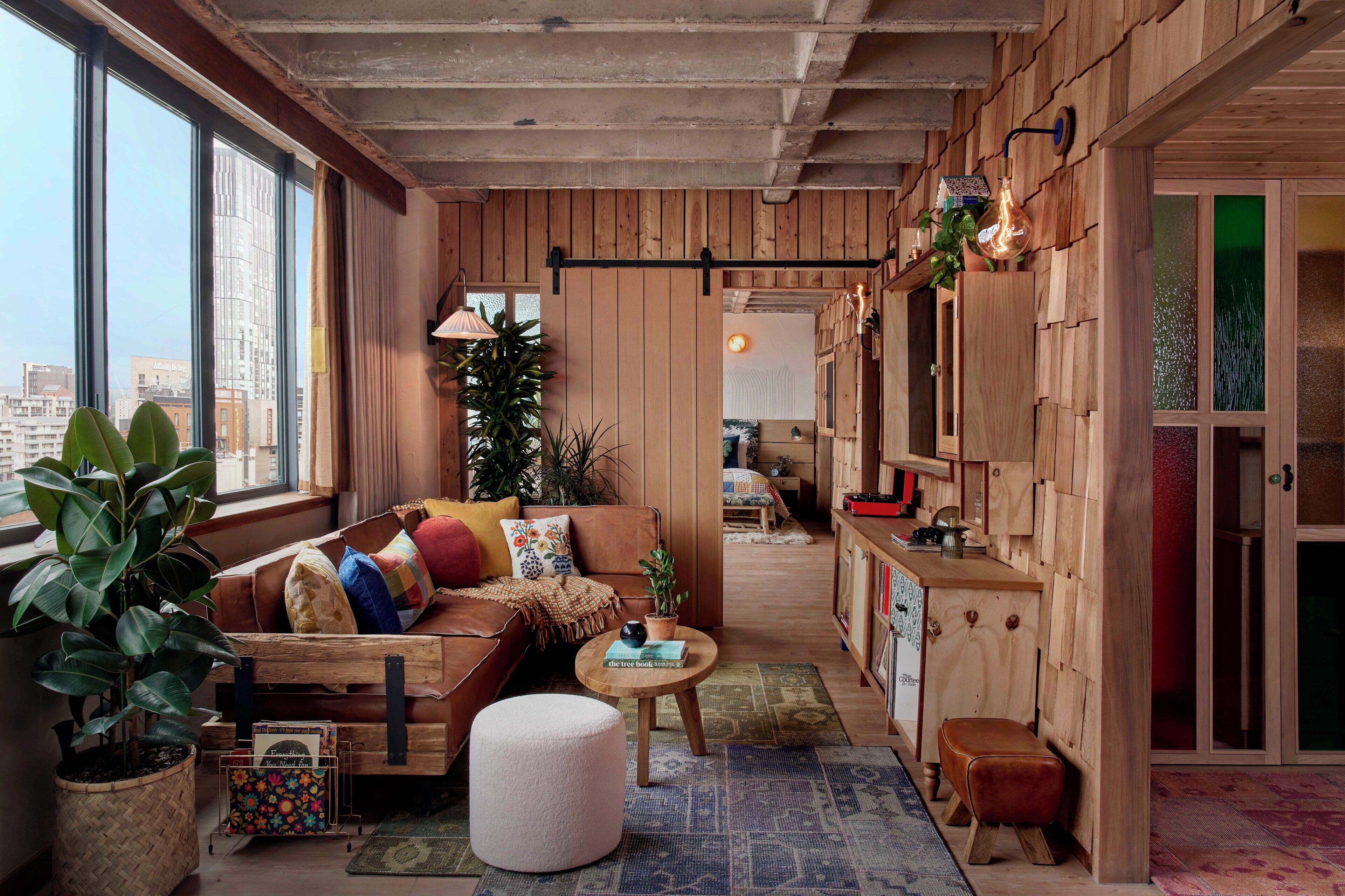 A cozy living room with a sofa, ottoman, and a coffee table, featuring a city view through large windows.