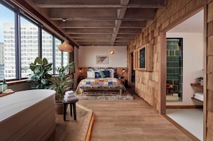 Un dormitorio con un ventanal grande, piso de madera, una cama con colcha colorida y vista a la ciudad.