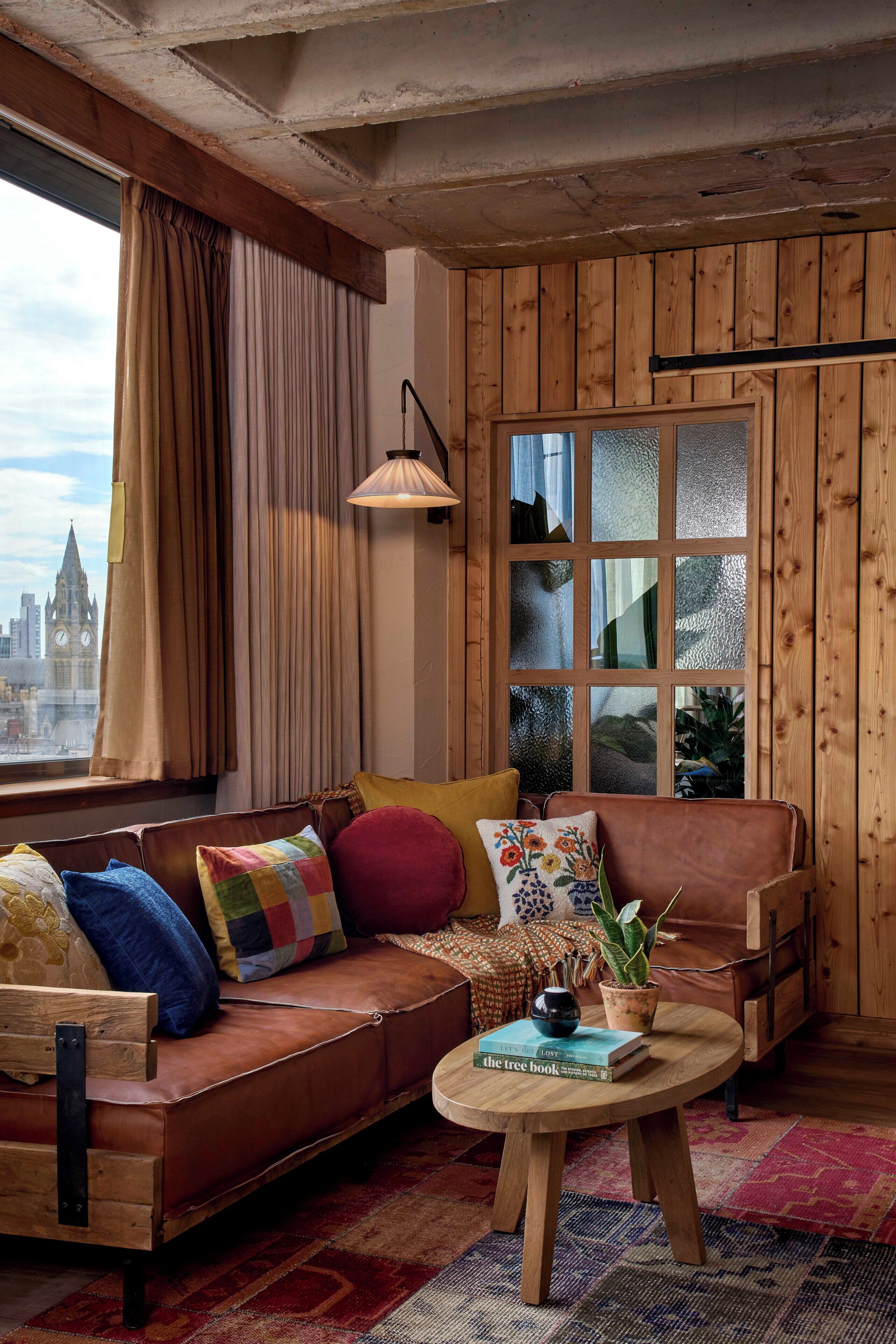 A cozy living room with a leather sofa, colorful cushions, a round wooden coffee table with a book and a plant, and a large window with a view of a cityscape.