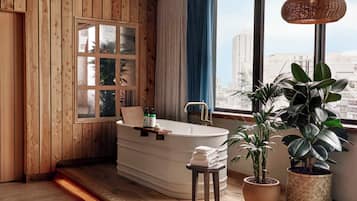 Un baño moderno con una bañera independiente, paneles de madera, un ventanal con vistas a edificios y plantas en macetas.
