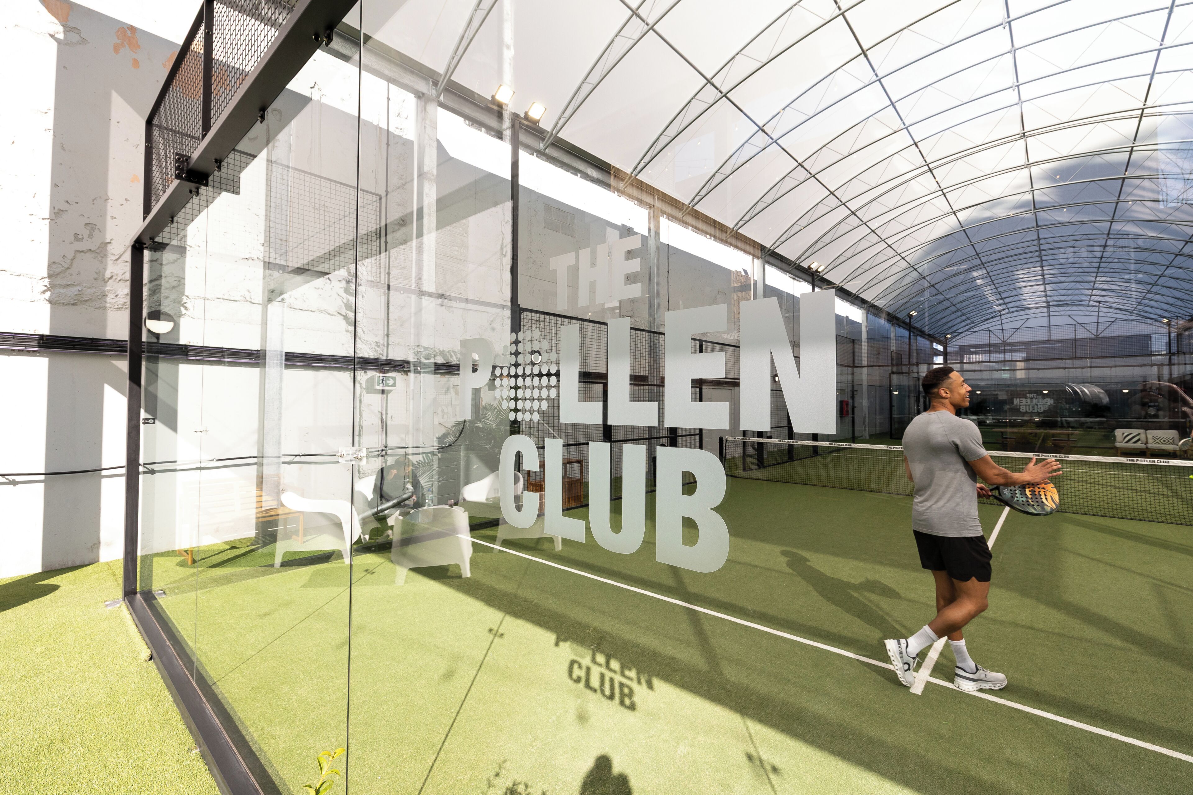 A tennis court with a net, a player in a grey shirt and black shorts, and a glass wall with 'The Bellen Club' text.