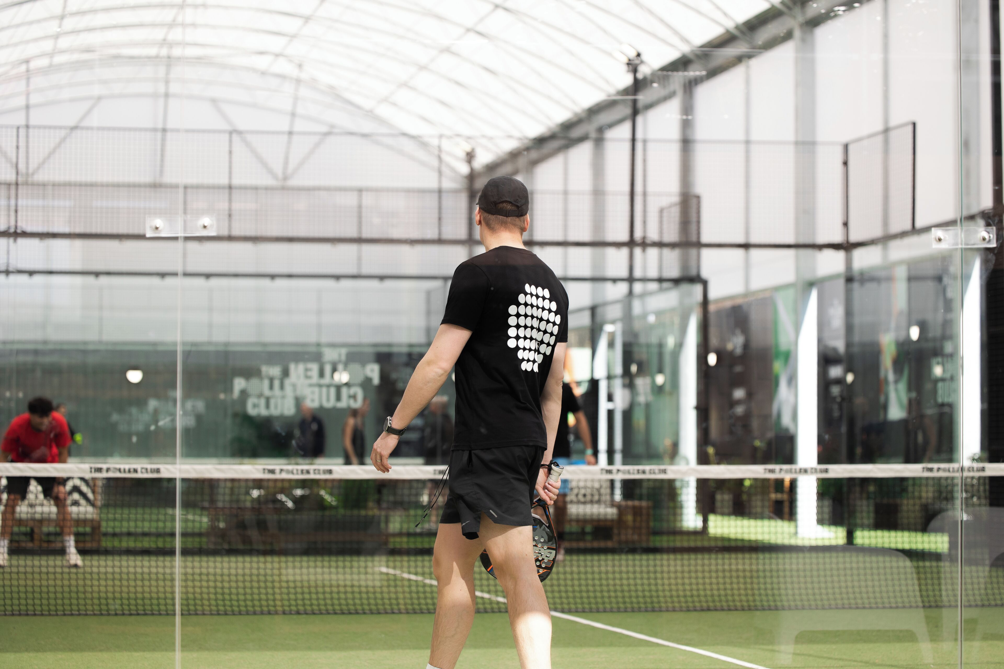 A tennis court with a net, a player in black attire, and a glass enclosure in the background.