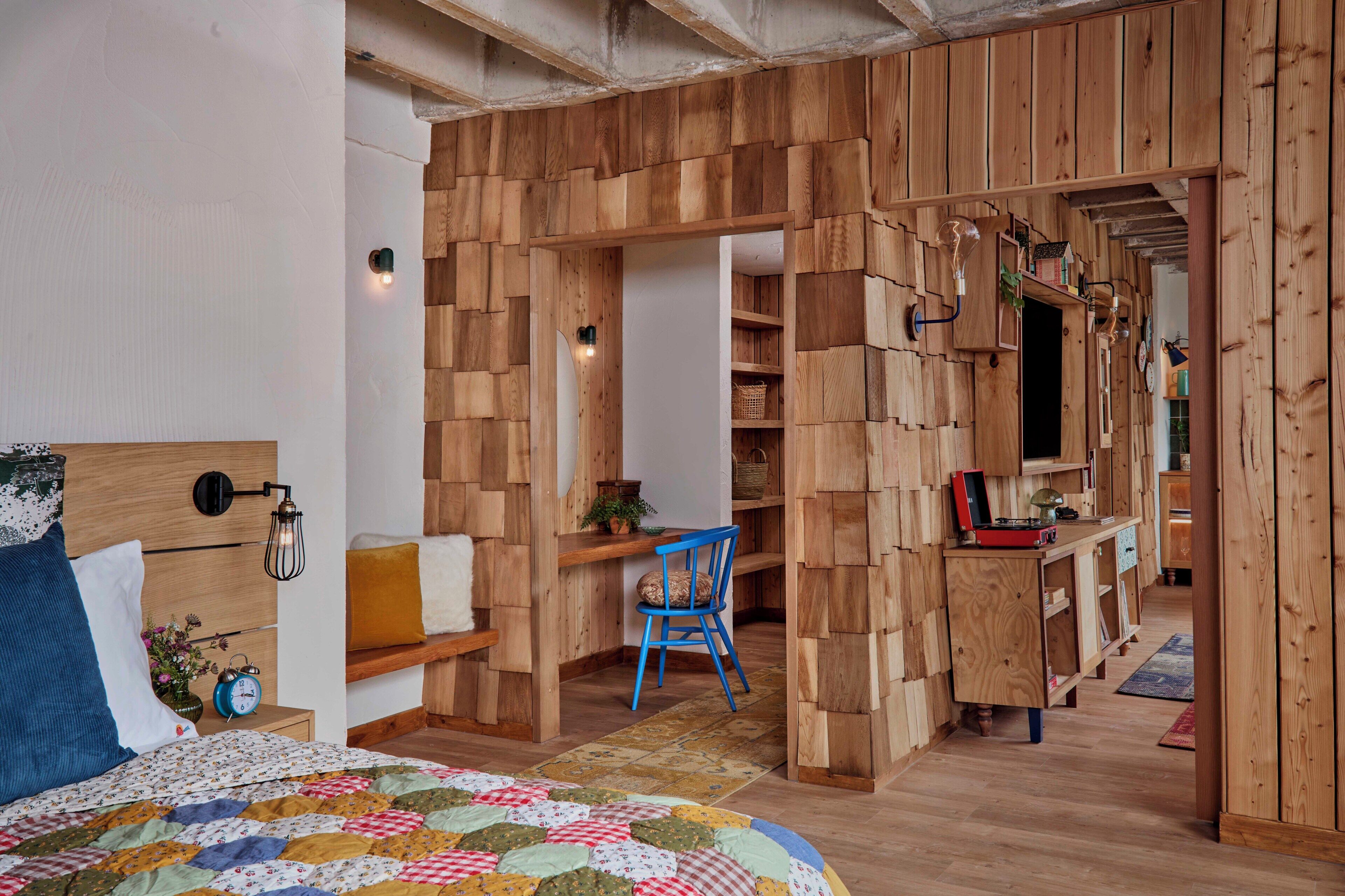 A room with a bed, a desk, and a chair, featuring wooden walls and a herringbone floor pattern.