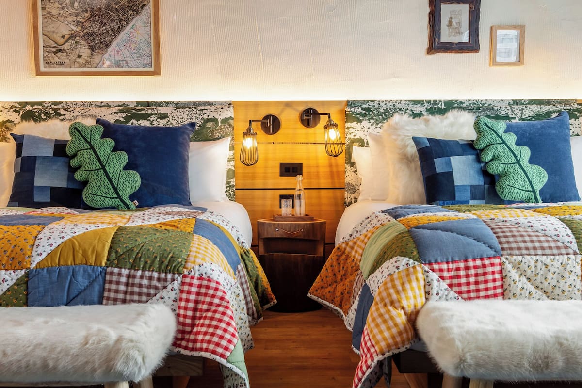 A room with two beds, a wooden bench, a patterned rug, and wall-mounted lighting.