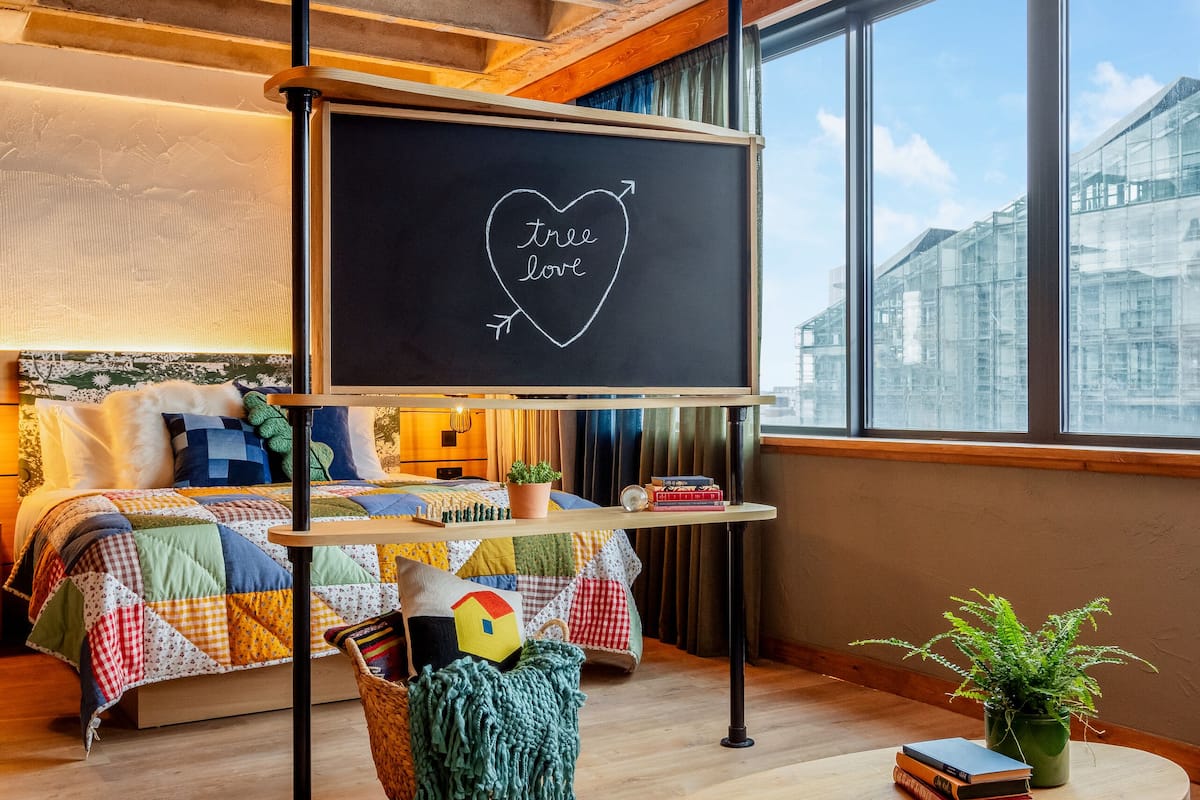 A cozy room with a bed, a round wooden table, a blackboard with 'tree love' written on it, and a view of buildings through large windows.