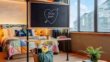 Una habitación acogedora con cama, una mesa redonda de madera, una pizarra con la inscripción 'tree love' y vistas a edificios a través de grandes ventanales.