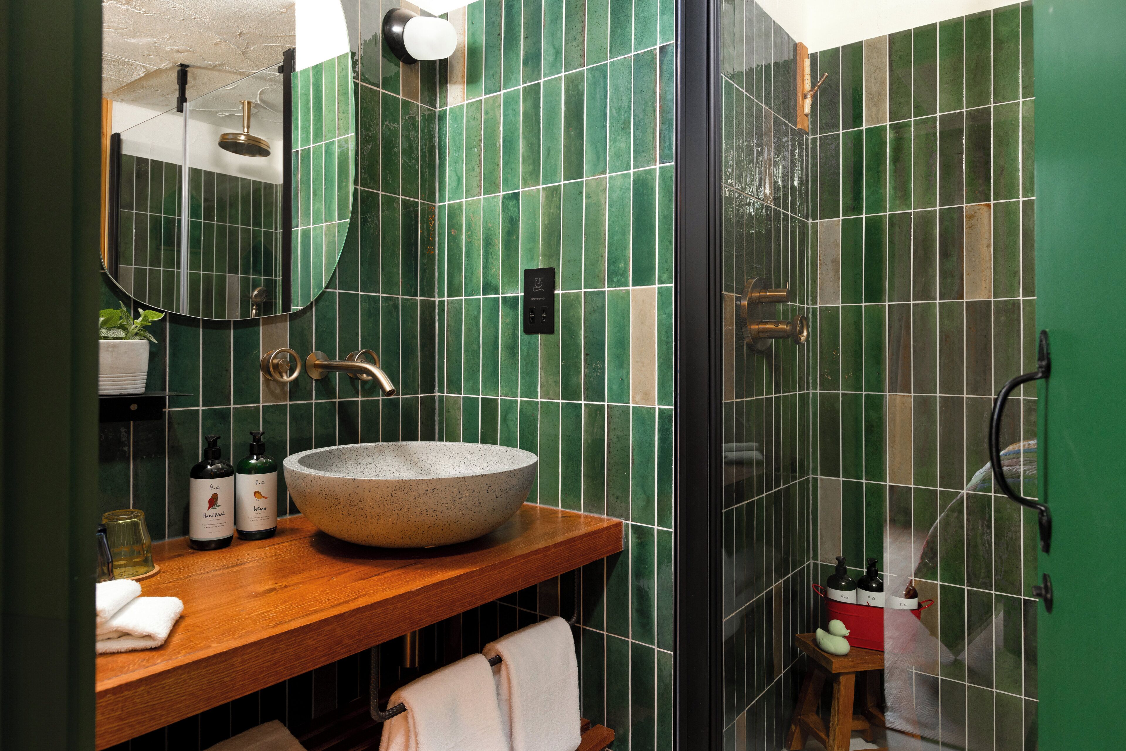 A bathroom with a wooden vanity, a large bowl sink, green tiled walls, a mirror, and a shower area.