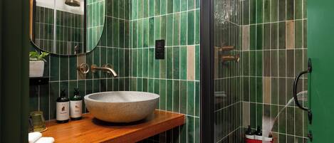 Un baño con tocador de madera, un lavamanos grande tipo cuenco, paredes cubiertas de azulejos verdes, un espejo y una zona de ducha.