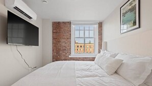 Une chambre avec un mur en briques, une fenêtre donnant sur la ville, un téléviseur à écran plat fixé au mur et un lit avec une literie blanche.