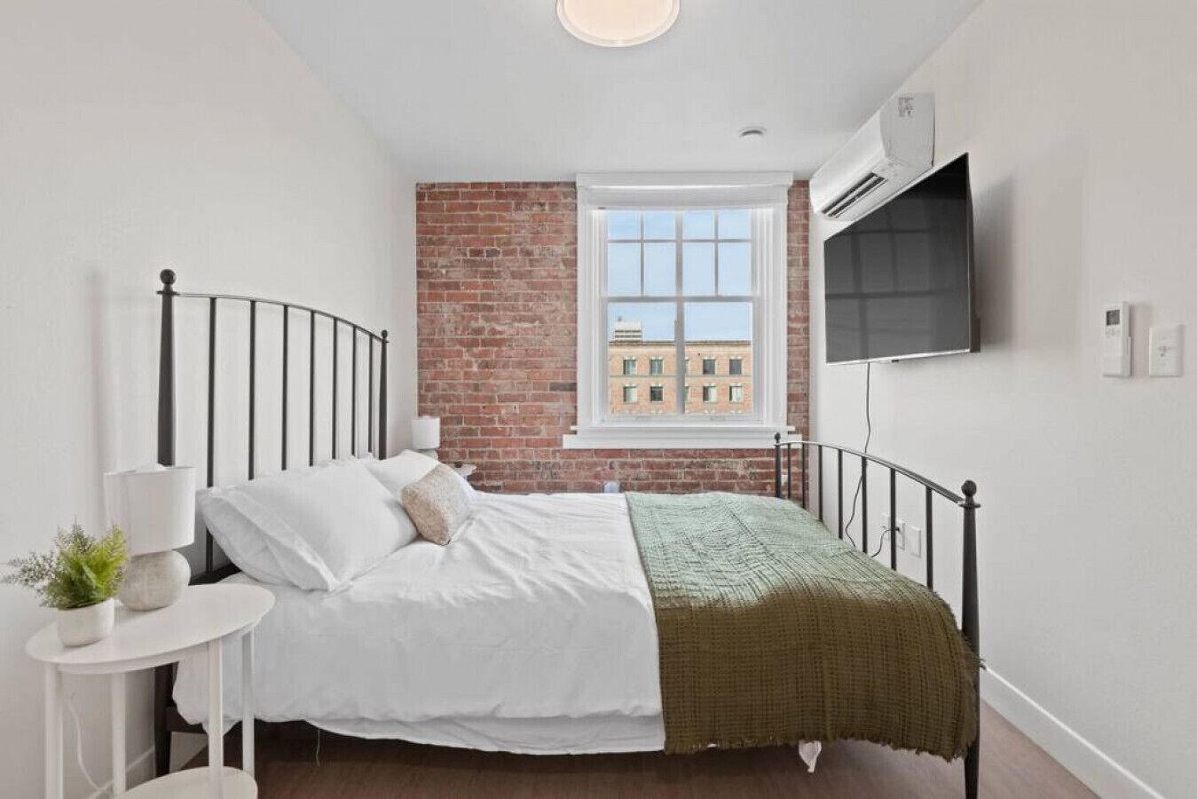 A bedroom with a brick wall, a window, a bed, a bedside table with a lamp, and a mounted TV.