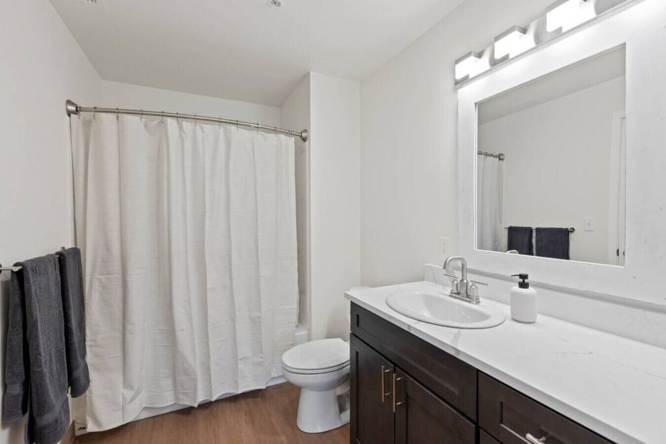 A bathroom with a white shower curtain, a toilet, a sink with a mirror above it, and dark wooden cabinets.